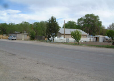 A paved street runs past a house with a fenced yard, several trees, and a parked van. Other small buildings and greenery are visible in the background under a cloudy sky.
