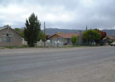 A small residential neighborhood with single-story houses, trees, a dirt road in the foreground, and hills visible in the background under a cloudy sky.