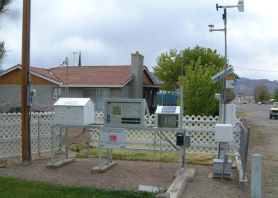 A weather monitoring station with multiple instruments and sensors is set up in front of a house, enclosed by a white fence.
