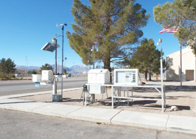 A small weather station with various instruments stands near a sidewalk and trees; mountains and an American flag are visible in the background.