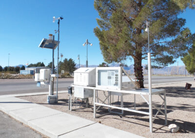 A meteorological station with instruments and sensors on metal tables is set up near a road, with trees and mountains in the background.