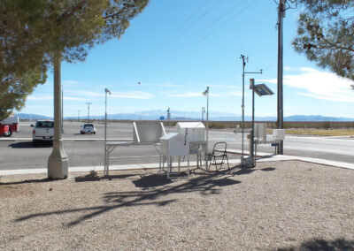 Outdoor weather monitoring station with various sensors and instruments, located near a paved parking lot and desert landscape, under a clear sky.