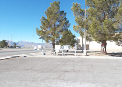 An empty parking lot with trees, utility boxes, and a white car partially visible on the right; mountains are seen in the background under a clear blue sky.