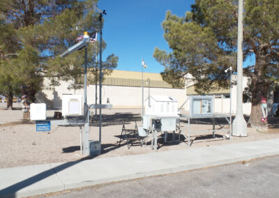 A weather station with various meteorological instruments and sensors is positioned on a gravel area near a building, surrounded by trees and a clear blue sky.