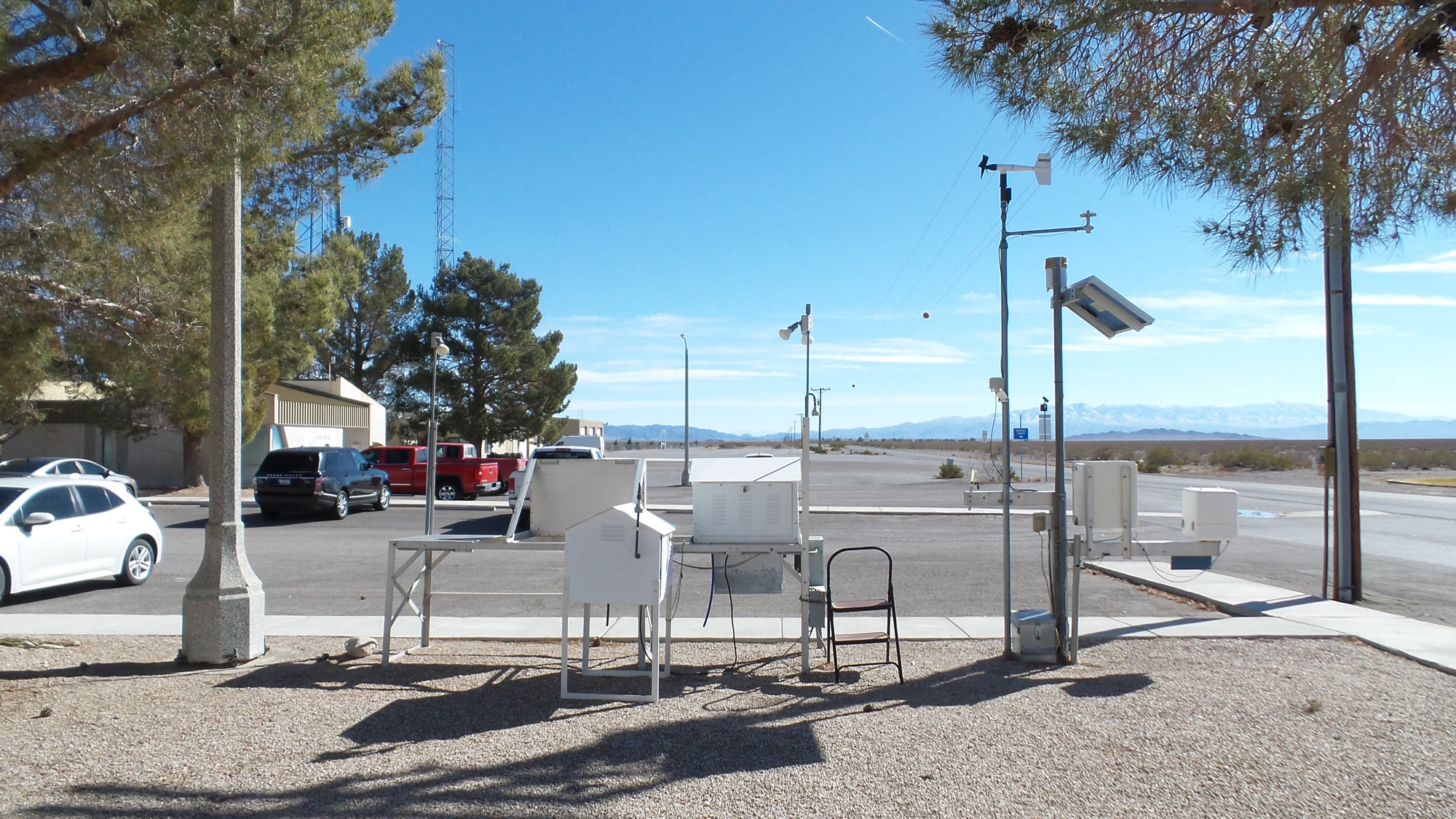 A weather station with instruments and sensors stands by a paved lot with parked cars, trees, and mountains visible in the distance under a clear blue sky.
