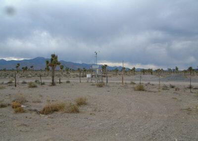 A weather monitoring station stands in a dry, desert landscape with sparse vegetation, surrounded by mountains under a cloudy sky.