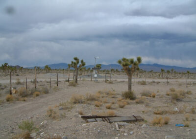 A weather station stands in a fenced area of a desert landscape with sparse vegetation and cloudy skies in the background.