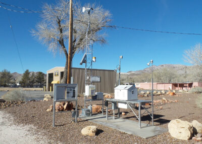 A small outdoor weather station with various sensors and equipment is set up on a concrete pad in a dry, rocky area with buildings, trees, and mountains in the background.