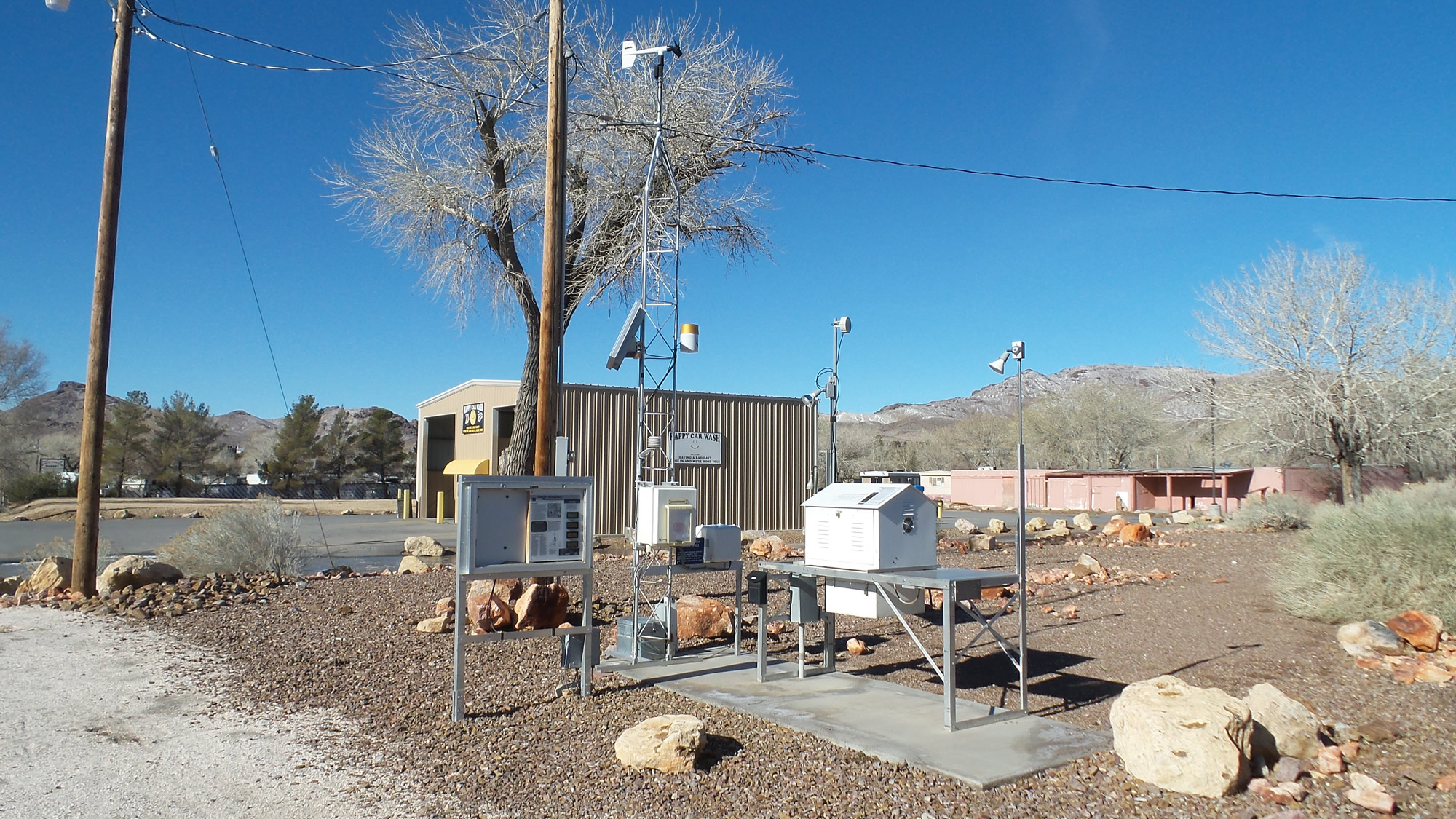 A small outdoor weather station with various sensors and equipment is set up on a concrete pad in a dry, rocky area with buildings, trees, and mountains in the background.