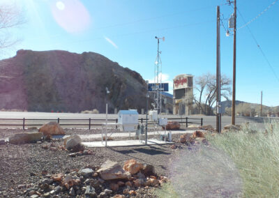 A roadside weather monitoring station with sensors and instruments stands near rocks and dry vegetation; a large rocky hill and water tower are in the background under a clear blue sky.