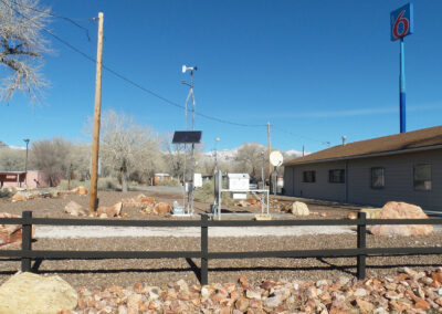 A weather monitoring station with solar panels and antennas stands near a one-story building under a clear blue sky; a Motel 6 sign is visible in the background.