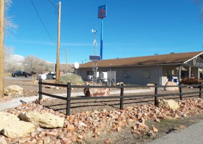 A single-story Motel 6 building with a large sign, surrounded by rocks and a fenced yard, under a clear blue sky.