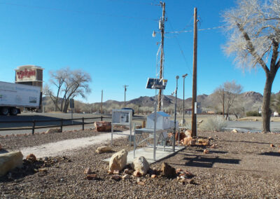 A white bench and mailbox sit on a gravel area near a road, with utility poles, a truck, trees, and mountains in the background under a clear blue sky.