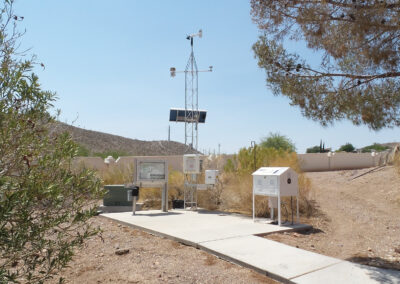 A small weather station with a solar panel and various instruments is set up on a concrete pad in a desert landscape with sparse vegetation.