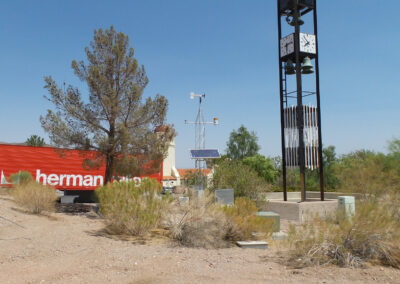 A desert landscape featuring a tall metal clock tower, sparse vegetation, utility boxes, and a red building with partial herman lettering visible.