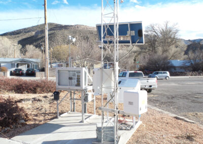 A weather monitoring station with solar panels and various instruments is installed on a concrete slab near a residential area with hills in the background.
