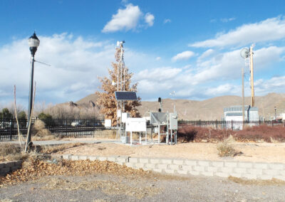A small weather monitoring station with solar panels stands on gravel surrounded by dry vegetation, with mountains and utility poles in the background under a partly cloudy sky.