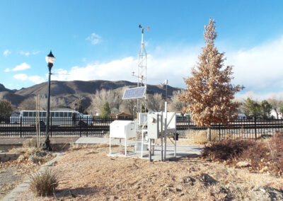 A weather monitoring station with solar panels and sensors stands near a tree and lamp post, with mountains and a fence in the background under a clear blue sky.