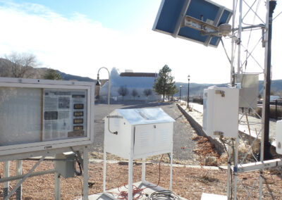 Weather monitoring equipment including display screens and sensors is set up outdoors on gravel with mountains and a building visible in the background.