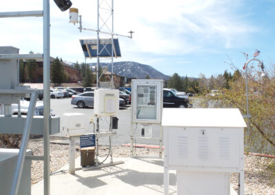 Weather monitoring station with various instruments and solar panel, set on a concrete pad next to a parking lot, with mountains and trees in the background.