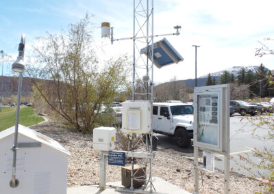 A weather monitoring station with various sensors and instruments, a display board, and a white vehicle parked nearby in a lot with mountains in the background.