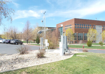 A weather station with sensors and a solar panel is installed on a landscaped area near a school building and parking lot under a partly cloudy sky.