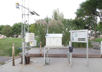 A weather monitoring station with instruments, a control box, solar panel, and informational sign, set on a concrete platform in a landscaped park area.