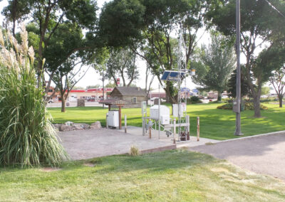 A weather monitoring station with various instruments sits on a concrete pad in a green park near trees and a grassy area, with buildings visible in the background.