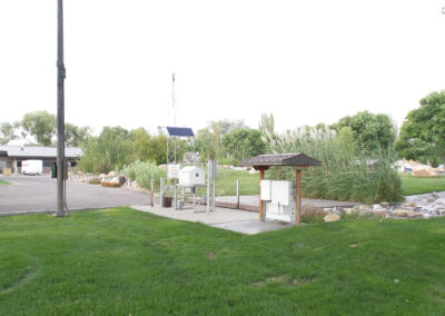 A small weather monitoring station with instruments and a solar panel sits on a concrete pad near grass, trees, and a parking area.