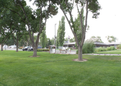A grassy park area with several tall trees, a flagpole with flags, a small structure, and parked vehicles near a low building in the background.