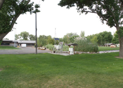 Open grassy area with trees, a paved path, informational sign, small fenced utility area, and buildings in the background under a cloudy sky.