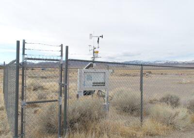 A fenced weather monitoring station with instruments including an anemometer and sensors, located in a dry, open field with mountains in the background.
