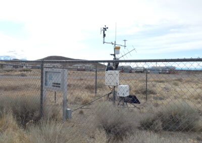A fenced-in weather station with various sensors and equipment stands in a dry, grassy field under a cloudy sky.