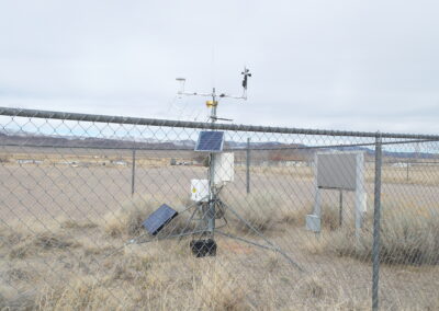 A weather monitoring station with solar panels and sensors is installed behind a chain-link fence in a dry, open landscape.