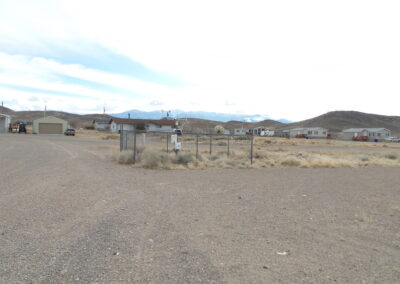 A fenced weather station stands on gravel near scattered houses, vehicles, and dry grass, with hills and mountains visible in the background under a cloudy sky.
