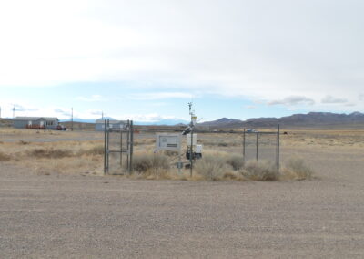 A small fenced weather station stands in a dry, open landscape with mountains in the background and a few scattered buildings nearby.