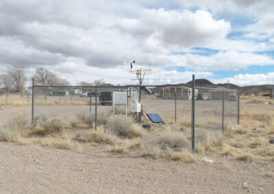 A fenced weather station with sensors and a solar panel is situated in a dry, grassy area, with buildings and mountains in the background under a cloudy sky.
