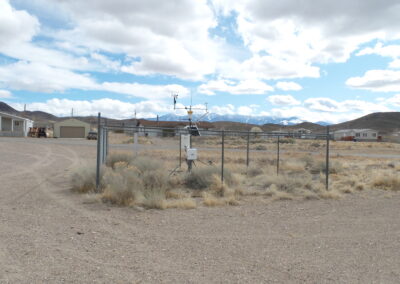 A fenced-in weather station with sensors is situated on dry, gravelly ground near residential buildings and mountains under a partly cloudy sky.