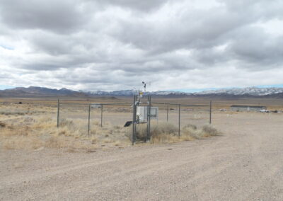 A fenced weather station stands in a dry, open landscape with mountains and cloudy sky in the background. Scattered buildings are visible in the distance.