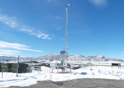 A weather monitoring station with a tall mast stands in a snow-covered fenced area, with mountains visible in the background under a clear blue sky.