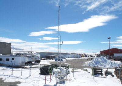 A fenced weather monitoring station with a tall metal tower stands in a snowy lot surrounded by industrial buildings and parked vehicles under a blue sky with scattered clouds.