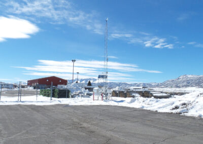 A fenced facility with a red building, radio tower, and snow-covered ground, set against a backdrop of mountains under a blue sky with scattered clouds.