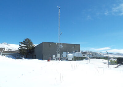 A fenced facility with antennas and equipment sits in a snowy landscape under a clear blue sky, with mountains visible in the background.