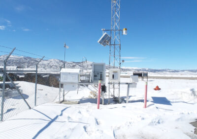 Weather monitoring station surrounded by snow, chain-link fence, and mountains in the background under a clear blue sky.
