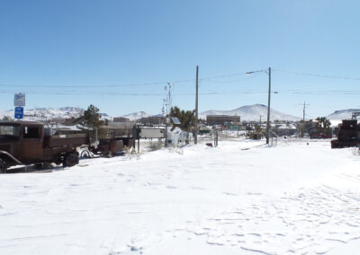 Old vehicles parked on a snow-covered lot in a small desert town, with mountains and buildings visible in the background under a clear sky.