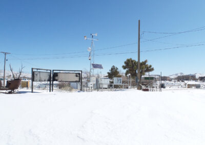 A snowy landscape with weather monitoring equipment, utility poles, and signs under a clear blue sky. Buildings and hills are visible in the background.