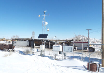 A weather monitoring station with instruments and solar panels stands in a snowy area near a fenced lot with trucks and equipment under a clear blue sky.