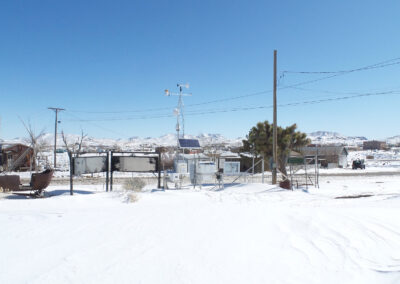 A snow-covered yard with scattered structures, utility poles, and a weather station. Distant mountains and a clear blue sky are visible in the background.