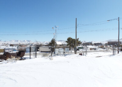 A snow-covered landscape with utility poles, sparse trees, and scattered buildings in the background under a clear blue sky.