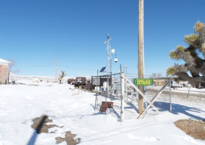 Snow-covered ground with various weather instruments inside a fenced area, a ENTRANCE sign, and a building and vehicles in the background under a clear blue sky.